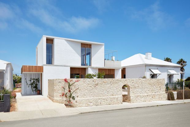 Street-facing view of a low-rise residential property combining a crisp white contemporary two-storey addition with a smaller white pitched-roof dwelling. The main house has clean rectangular volumes, tall vertical glazing, a timber-clad garage or entry element, and a rooftop or upper-level terrace with a glass balustrade. A rough pale stone boundary wall and simple front path define the streetscape, with sparse coastal-style planting under a bright blue sky.