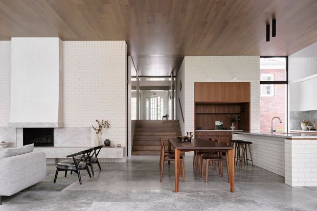 Open-plan contemporary kitchen, dining and living area with a polished concrete floor, white brick-tile walls, extensive timber ceiling lining, and a warm timber built-in dining and kitchen feature. The room includes a dining table with chairs, kitchen island and stools, sculptural white fireplace surround, staircase, and large windows.