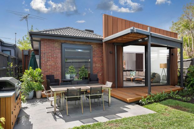 A contemporary single-storey residential extension viewed from the rear garden. The original red-brick section has a dark tiled roof, while the addition features vertical timber cladding, a projecting flat-roofed pergola-like structure, extensive glazing, and a timber deck. A paved outdoor dining area and built-in outdoor kitchen/barbecue sit in the foreground, with the living room visible through the glazed doors.