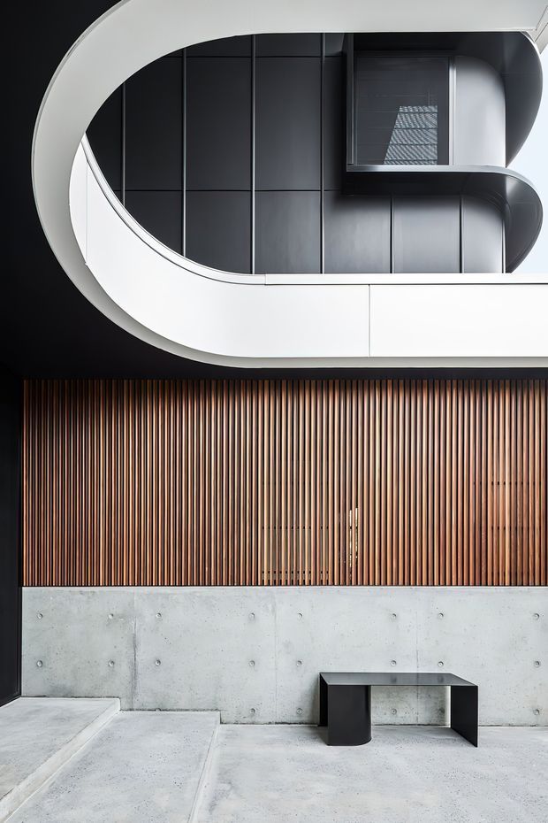 A refined contemporary building facade with a curved white projecting balcony or canopy above a dark recessed upper level. The lower facade combines vertically slatted timber screening with exposed light concrete walls and paving. A simple black metal bench sits in the foreground, while the upper glazing reflects a gridded structure. The composition emphasizes strong geometric curves, vertical rhythm, and contrasting black, white, timber, and concrete surfaces.