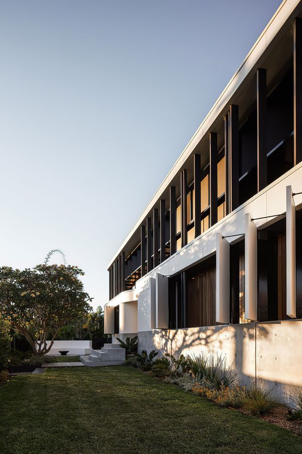 Exterior architectural photograph of a contemporary multi-storey building viewed from a landscaped garden. The facade combines pale concrete or rendered wall surfaces with dark vertical columns, recessed glazing, timber-toned screening and a strong linear roof edge. A sculptural curved white entry element, steps, lawn and planted garden soften the rectilinear structure. Warm low-angle sunlight creates strong shadows across the facade.