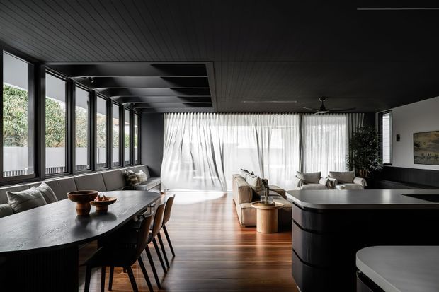 A sophisticated open-plan interior combining a dining area, living room, and kitchen. The space features extensive black-framed glazing along one side, sheer white curtains, dark timber-lined ceiling, warm timber flooring, a large dark dining table, and a sculptural black kitchen island. The restrained palette and clean lines create a refined, dramatic contemporary atmosphere.