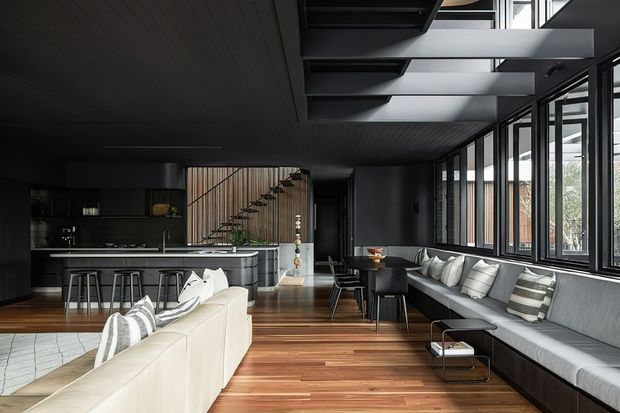 A dark, sophisticated open-plan living space with a kitchen at the rear, a long dining area, and a large lounge with built-in window seating. The room features extensive black-framed glazing, exposed dark structural beams and a high-level glazed roof section, a timber-lined stair screen, charcoal cabinetry, a pale sectional sofa, and warm timber flooring.