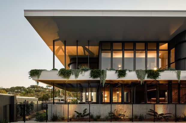 Contemporary multi-level residential building viewed from the exterior at dusk, featuring broad flat roof planes, extensive dark-framed glazing, projecting balconies, slim supports, and cascading greenery along the upper terrace. Warm interior lighting contrasts with the dark facade, while a metal picket fence and landscaped frontage sit in the foreground.