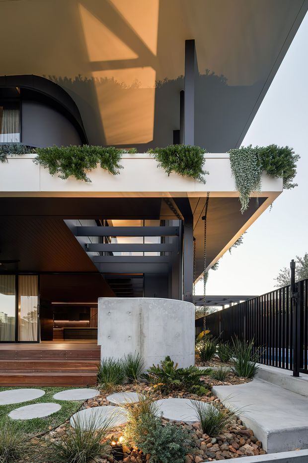 A contemporary multi-level residential exterior courtyard or entry approach, viewed from outdoors. The composition features a dramatic cantilevered upper floor with a planted balcony edge, dark timber soffits, exposed concrete, black metal screening and fencing, broad timber steps, and a sculptural drought-tolerant garden with gravel, grasses and circular concrete stepping pads. Warm interior lighting glows beneath the overhang, while a partially visible curved architectural element appears at upper left.