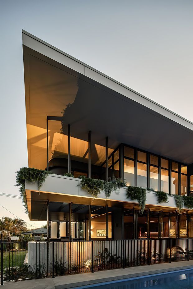 Exterior architectural photograph of a contemporary multi-storey residence at dusk. The building has a strong flat roof with broad overhangs, extensive dark-framed glazing, projecting balconies with cascading planter greenery, warm interior lighting, and a black metal boundary fence. A swimming pool occupies the foreground.
