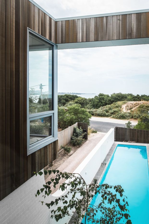 Exterior architectural view of a contemporary coastal residence, showing dark vertical timber cladding, large aluminium-framed glazing, a projecting upper-level connection, and a narrow swimming pool beside the house. Low coastal vegetation, a sandy access path, and the ocean are visible beyond the property.