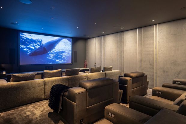 A dark, luxurious home cinema with multiple rows of large upholstered reclining seats facing a wall-mounted projection screen. The room features subdued recessed ceiling lighting, acoustic-style vertical wall panels in a warm grey finish, dark carpeting, and a blue-lit underwater image displayed on the screen.
