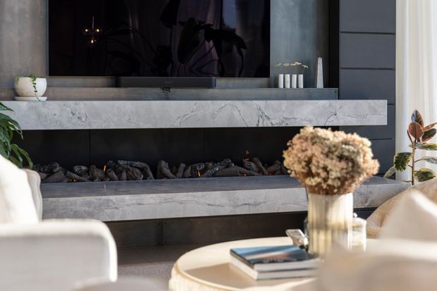 A refined contemporary living room centered on a linear fireplace with layered pale marble or stone mantels and a dark firebox. A large television is mounted above the fireplace, with a round light-toned coffee table, books, vase, and upholstered seating in the foreground. Tall curtains and indoor plants frame the space, creating a polished, warm-neutral interior.