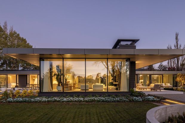 Twilight exterior view of a low, single-storey contemporary residence with broad flat roof planes, extensive floor-to-ceiling glazing, warm interior lighting, and a central glazed living space. Landscaped lawn, garden beds, paved paths, and a curved foreground wall frame the house. A raised roof structure or skylight-like element is visible above the central volume.