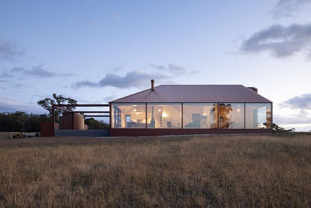 Single-storey contemporary house photographed from the surrounding rural landscape at dusk. The long rectangular building has extensive floor-to-ceiling glazing, a simple pitched metal roof, dark slender window frames, and a covered open-sided carport or terrace structure extending to the left. Warm interior lighting reveals living spaces inside, while dry grassland and scattered trees form the setting.