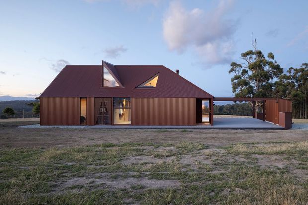 Single-storey contemporary rural house with a rust-red corrugated metal-clad facade and pitched roof, punctuated by angular dormer-style skylights and tall glazed openings. A covered carport extends from the right side, with open grassland and trees surrounding the building.