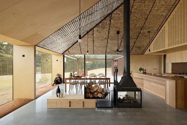 Open-plan contemporary living and dining space with a warm timber-lined ceiling, full-height glazing, polished concrete flooring, and extensive light timber cabinetry. A central black freestanding fireplace creates a strong focal point between the lounge and dining areas, while the dining zone opens toward a landscaped exterior.