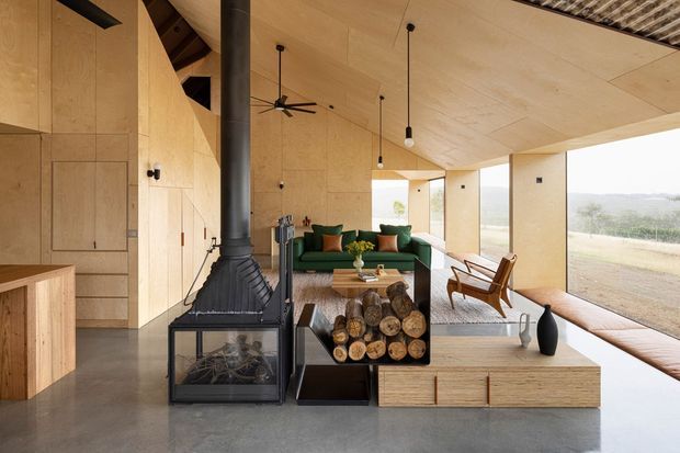 Warm contemporary open-plan living space with full-height pale timber wall and ceiling lining, polished concrete flooring, a black freestanding wood-burning fireplace, and large glazed openings overlooking a rural landscape. A deep green sofa, timber coffee table, leather lounge chairs, and integrated timber storage create a relaxed, refined interior. Exposed roof structure and simple pendant lights add architectural character.