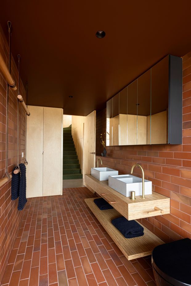 A warm, narrow bathroom with exposed reddish-brown brick walls and matching brick floor tiles. A long timber vanity supports two white rectangular vessel basins with brushed brass gooseneck taps, beneath a large mirrored cabinet. Pale timber cabinetry conceals storage on the left, while a carpeted stairway leads upward at the rear. The dark brown ceiling, warm lighting, navy towels, and timber joinery create a rich, earthy interior.