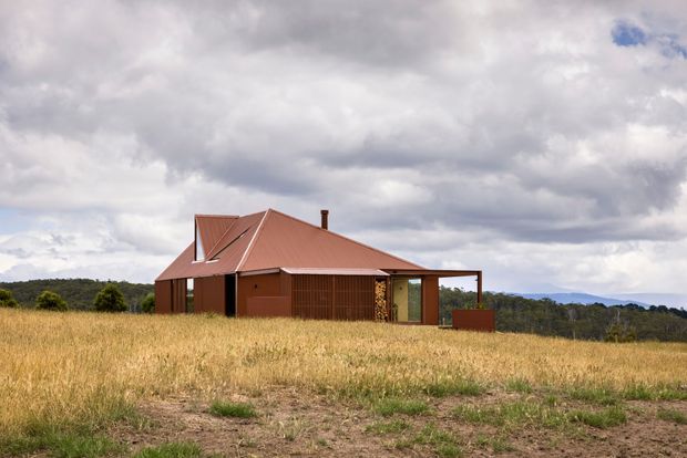 A compact single-storey rural house set in a dry grass field, photographed from the exterior. The building has a distinctive steep corrugated metal gable roof in a muted terracotta-red finish, matching dark red-brown wall cladding, large glazed openings, and a simple covered outdoor area. Rolling wooded terrain and a dramatic overcast sky form the background.