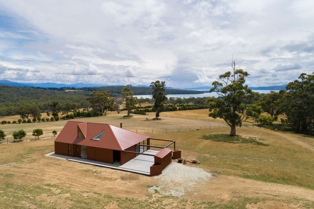 Aerial view of a compact single-storey rural house set within open dry farmland, with a distinctive red pitched metal roof, dark reddish-brown exterior walls, a rooftop skylight, and a broad pale deck or terrace. Mature trees, fields, distant hills, and a large body of water form the surrounding landscape.