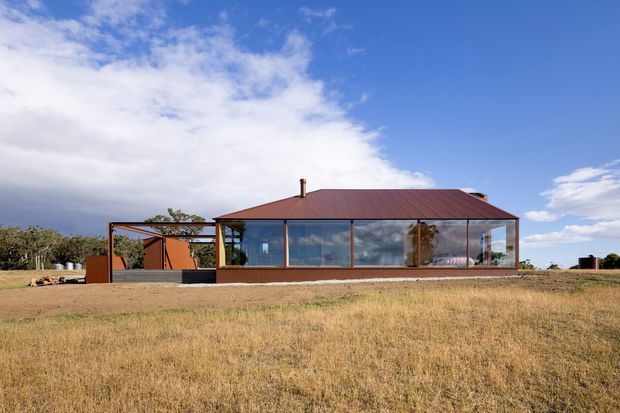 Single-storey contemporary rural residence viewed from across a dry grass field. The house has extensive floor-to-ceiling glazing framed in dark metal, a low-pitched corrugated metal roof, a red-brown solid wall section, and a covered open-sided outdoor structure extending from the left. The surrounding open field and distant vegetation create a spacious rural setting under a partly cloudy blue sky.
