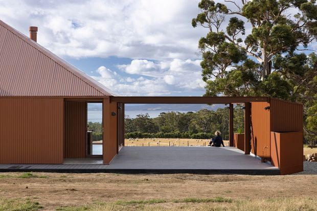 Exterior view of a low, barn-like rural building with a steep corrugated metal gable roof and warm rust-brown vertically ribbed cladding. A large central glazed opening and covered outdoor terrace frame views across open pasture and eucalyptus trees. The composition highlights the building's simple rectangular form, strong indoor-outdoor connection, and broad timber-toned deck.