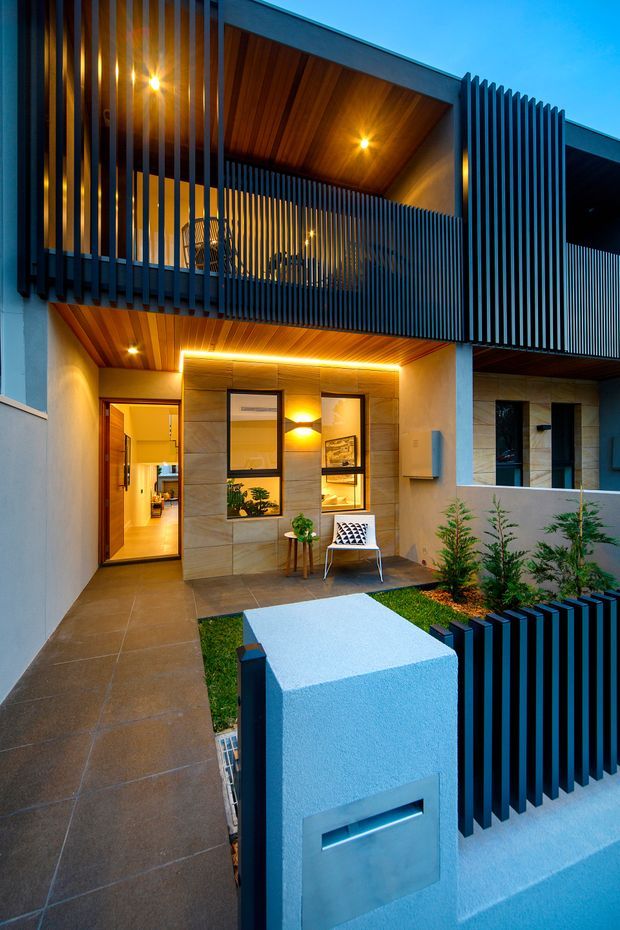 Twilight exterior view of a contemporary multi-storey residence with a recessed entry courtyard. The facade combines pale rendered walls, warm timber-lined soffits, stone-look wall panels, and prominent dark vertical metal screening around the upper balcony and front boundary. A tiled walkway leads to a glazed timber entry door, with a small outdoor chair and table beside the entrance, landscaped planting, and integrated warm lighting.