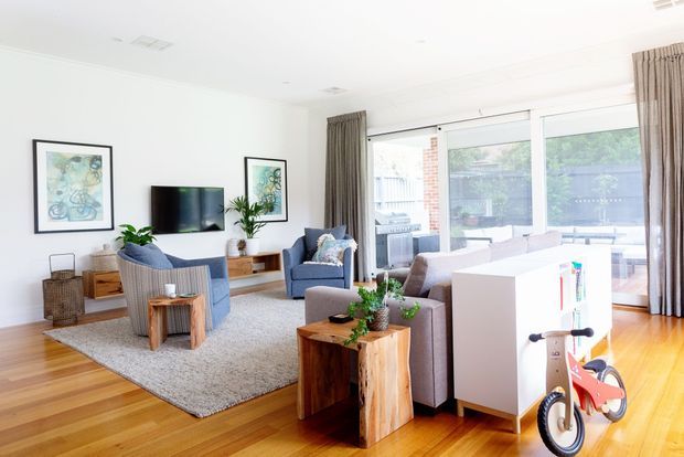 Bright, open-plan living room with timber flooring, a large textured area rug, blue upholstered lounge chairs, a grey sofa, timber coffee and side tables, wall-mounted television, framed artwork, and indoor plants. Full-height glazed doors and windows open toward an outdoor area, with neutral curtains and abundant natural light. A white storage unit and children's bicycle appear in the foreground.