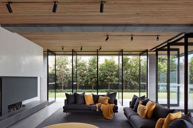 A contemporary living room with expansive floor-to-ceiling glazed walls opening toward a landscaped garden. The room features dark charcoal sectional sofas with mustard and ochre cushions and throws, a linear built-in fireplace set into a pale wall, a grey carpet, extensive timber slatted ceiling lining, black-framed glazing, and track lighting. The space is bright, open, and refined, with a strong indoor–outdoor connection.