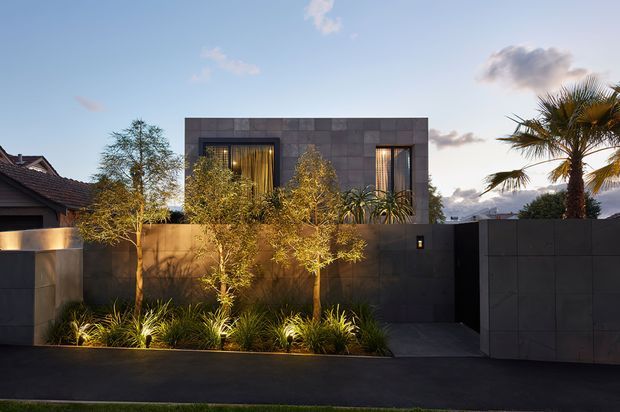 Contemporary residential facade viewed from the street at dusk, with a dark charcoal rendered or panelled upper volume, recessed illuminated windows, a solid boundary wall and pedestrian entry gate. Integrated landscape lighting highlights a narrow garden bed with ornamental grasses and small trees, while a palm rises behind the wall. Neighbouring rooflines are visible at the edges.