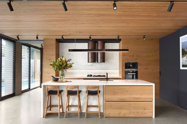 Contemporary open-plan kitchen dominated by a large light-wood island with three bar stools, integrated drawers, a pale stone benchtop, and a cooking area against a pale stone feature splashback. The room has extensive louvred glazing on the left, a warm timber slatted ceiling, timber wall panelling, black track lighting and dark charcoal accents. A framed landscape image is visible on the right wall.