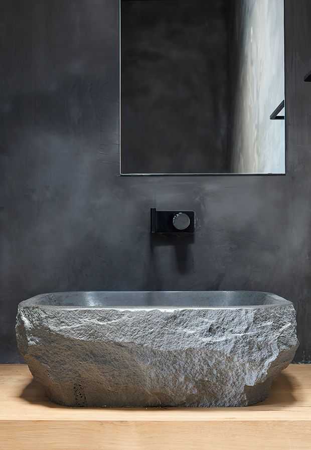 A moody minimalist bathroom featuring a rough-hewn natural stone freestanding bathtub or basin-like tub against a dark textured plaster wall. A rectangular mirror, dark wall-mounted tapware, and warm timber flooring create a stark contrast between the charcoal wall, pale stone fixture, and honey-toned wood.