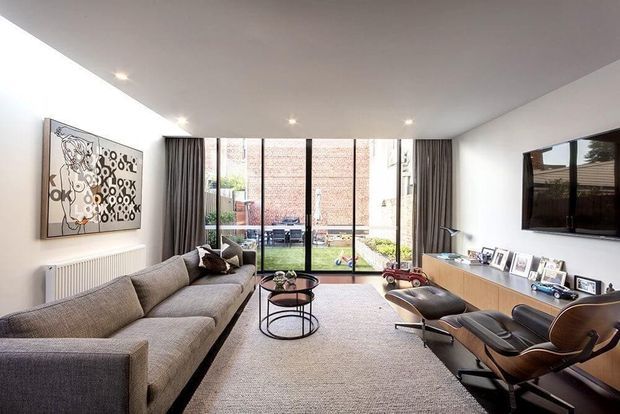 Contemporary living room with a large L-shaped grey sectional sofa, mid-century-style lounge chairs, round nesting coffee tables, a long timber media console, wall-mounted television, framed artwork, and floor-to-ceiling glazed doors opening to a small courtyard. Neutral finishes, recessed ceiling lights, and full-height dark curtains create a refined, softly lit interior.