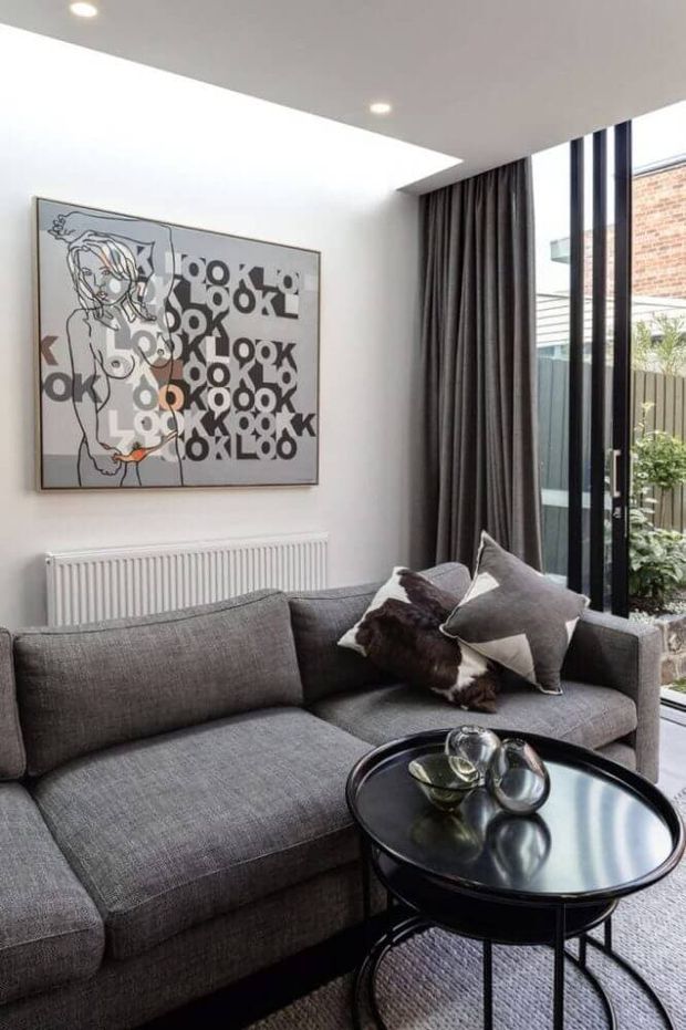 A compact contemporary living room with a large charcoal-grey sectional sofa, round black coffee table, monochrome wall artwork, and dark full-height curtains beside black-framed glazed doors opening to a small outdoor area. Recessed ceiling lights, white walls, and a radiator create a clean, modern interior with a strong grey, black, and white palette.