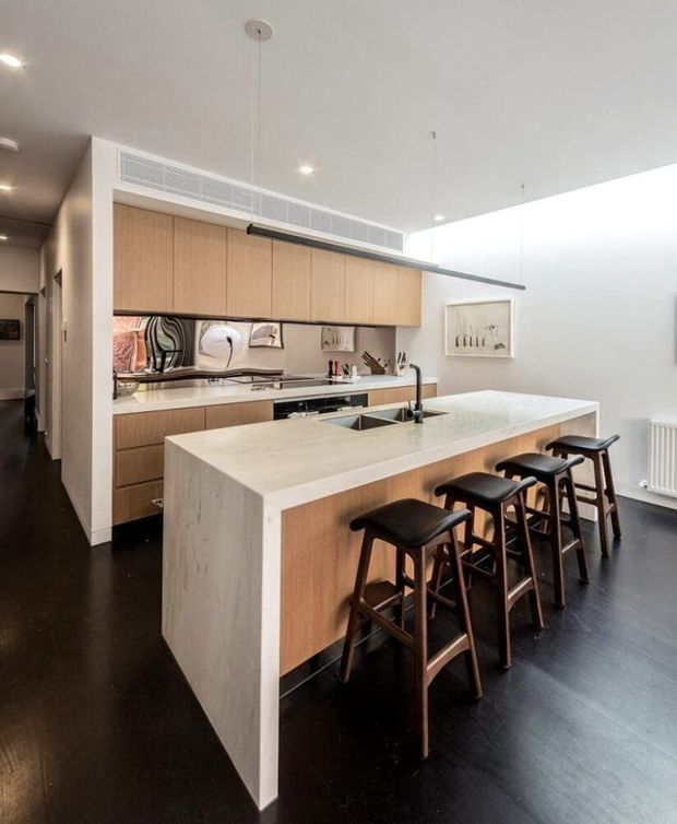 A bright contemporary kitchen viewed from the adjoining hallway, featuring a large white stone island with integrated sink and seating for four, warm timber cabinetry, a dark floor, stainless-steel appliances and a simple linear pendant light. White walls and ceiling create a clean, refined appearance, while artwork and sculptural backsplash details add subtle character.