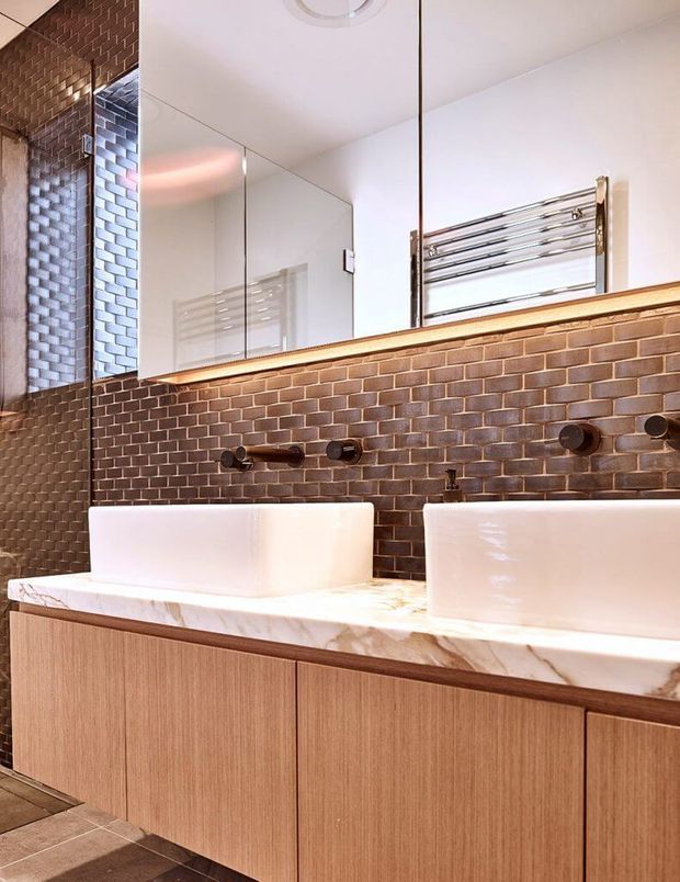 A contemporary bathroom vanity featuring twin white vessel basins on a marble-look benchtop, a warm timber floating cabinet, and a dark brown brick-pattern tiled splashback and wall. Large mirrored cabinetry reflects the room, while wall-mounted dark tapware and a polished chrome towel rail provide functional details. The space has warm layered lighting and a refined, high-contrast material palette.