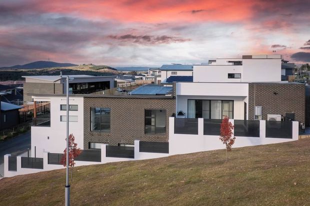 Exterior view of a contemporary multi-level residential development on a sloped suburban site. The buildings feature flat roofs, crisp white rendered volumes, dark brown brick veneer, large rectangular windows, dark metal privacy screens and balconies. A landscaped grass verge and street lighting sit in the foreground, with hills and a dramatic sunset sky beyond.
