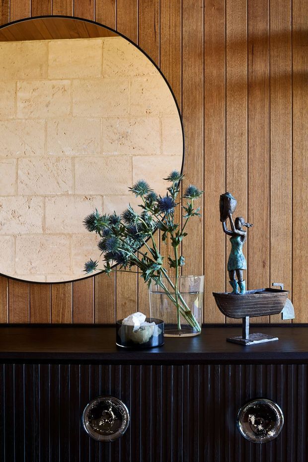 Close-up interior vignette featuring a dark timber console with fluted drawer fronts and round metallic pulls. Behind it is a warm mid-toned vertical timber-panelled wall with a large irregular rounded-edge light stone or plaster feature panel. The console displays a glass vase of blue thistles and a sculptural metal artwork depicting a figure in a boat. The composition is warm, textural, refined, and strongly influenced by mid-century styling.