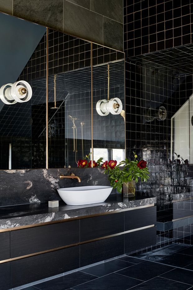 Dramatic dark bathroom interior featuring black grid-pattern wall tiles, large mirrored panels with brass-toned trim, a white vessel basin, dark marble vanity benchtop, black wall-mounted cabinetry, and a sculptural white wall light. A shower area is visible in reflection and to the right. Red flowers provide a small contrasting accent.