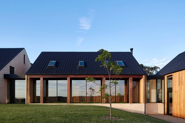 A contemporary single-storey residential building viewed from a landscaped lawn. The central pavilion has a steep dark metal gable roof with several skylights, extensive floor-to-ceiling glazing, and a regular rhythm of warm timber structural posts and framing. Adjacent pale rendered and timber-clad wings connect on either side, creating a courtyard-like composition. The image is captured in bright daylight with a clear blue sky and distant landscape visible through the glazing.