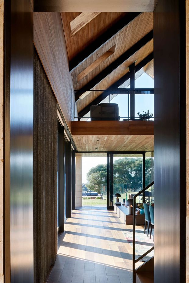 Interior view through a dark-framed opening into a bright double-height living and dining space. The room features a dramatic pitched timber-lined ceiling with exposed dark structural beams, a timber-clad mezzanine or upper level, extensive floor-to-ceiling glazing, and a tiled floor. Contemporary furniture and dining seating are visible beside the glazed garden outlook.