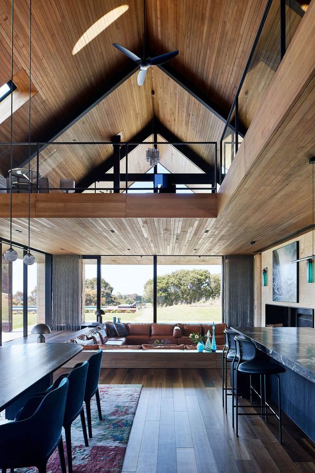 A dramatic double-height open-plan living, dining and kitchen interior with a prominent timber-lined vaulted ceiling and mezzanine level. Black-framed glazing opens the room to a green outdoor setting. The space includes a large sectional sofa, dining table, dark kitchen island with bar seating, pendant lights, ceiling fan, and dark metal balustrades. The design combines warm timber surfaces with charcoal finishes, leather seating, and abundant natural light.
