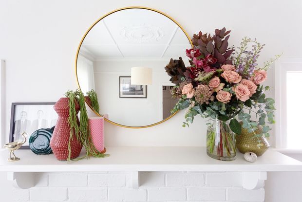 Bright, styled interior vignette of a white-painted brick fireplace mantel. The mantel displays framed artwork, decorative glass vessels, textured sculptural objects, and a large floral arrangement in a clear vase. A large round mirror with a thin brass-gold frame hangs above, reflecting a light-filled room with an ornate ceiling detail and table lamp.