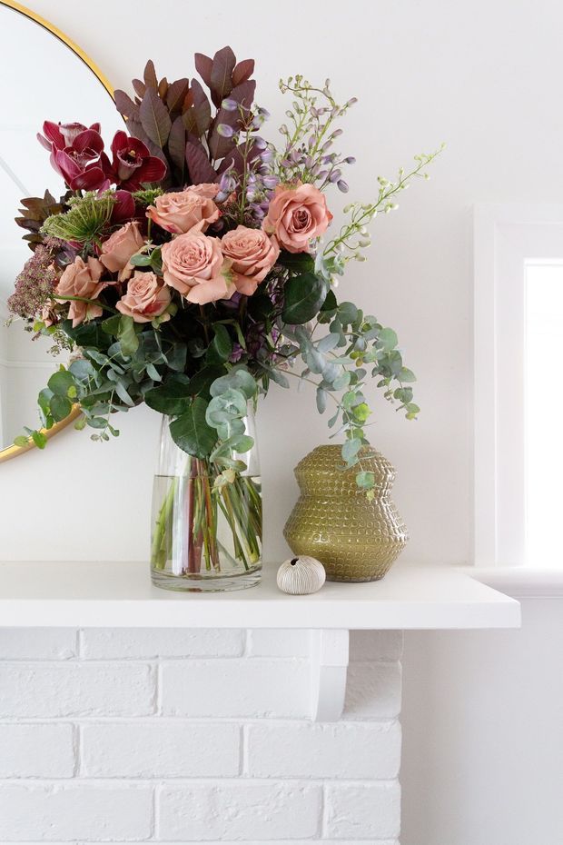 A close-up interior vignette featuring a large, colourful floral arrangement in a clear glass vase displayed on a white-painted brick fireplace mantel. A textured amber glass vessel and small decorative object sit beside it, with a partial round mirror visible against the white wall.