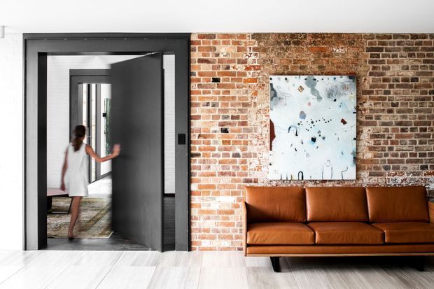 A contemporary interior featuring an exposed red-brick feature wall, a large dark-framed pivot door opening toward a hallway or adjoining room, and a cognac-brown leather sofa beneath abstract artwork. Pale timber flooring and white walls create contrast with the brick and dark door frame.