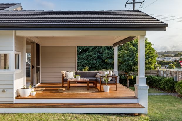 Exterior view of a single-storey home with a covered timber front veranda, white-painted posts and trim, horizontal weatherboard cladding, and a dark tiled pitched roof. The veranda contains outdoor lounge furniture and overlooks a landscaped suburban setting.