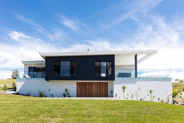Contemporary two-level residence viewed from the front, featuring a flat roof with broad overhangs, crisp white rendered walls, a dark vertically profiled facade section, expansive glazing, a timber garage door, and glass balcony balustrades. The home is set within a landscaped lawn under a bright blue sky.