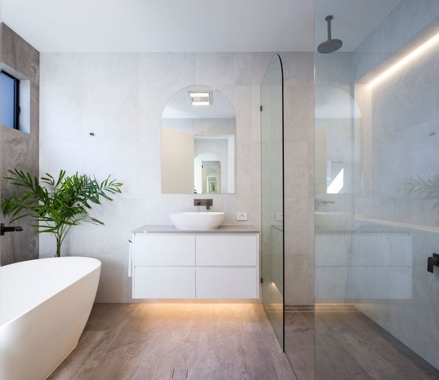 Bright contemporary bathroom featuring a freestanding white bathtub, wall-mounted white vanity with integrated under-lighting, vessel basin, arched mirror, large pale stone-look wall and floor finishes, and a frameless glass walk-in shower. A tall indoor plant adds a natural accent, while recessed and concealed lighting create a clean, refined atmosphere.