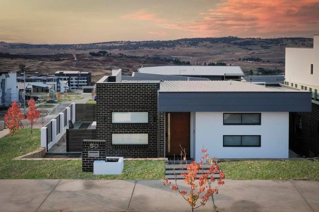Street-facing view of a contemporary low-rise house in a planned suburban development, featuring a dark brick rectangular volume, white rendered walls, broad flat roof forms, horizontal windows, and a landscaped front setback. Rolling open hills and neighboring modern buildings are visible beyond the property.