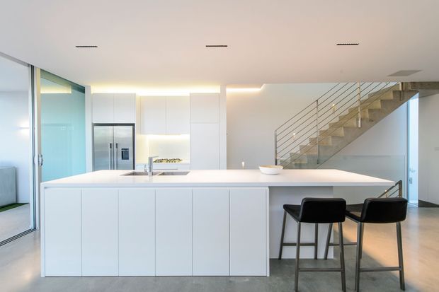 Bright, minimalist contemporary kitchen with a large white island and integrated cabinetry, stainless-steel refrigerator, subtle under-cabinet lighting, and two dark bar stools. A glazed sliding door is visible at left, while an open-riser concrete staircase with slim metal balustrading extends through the background. Pale polished flooring and white surfaces create a clean, airy interior.