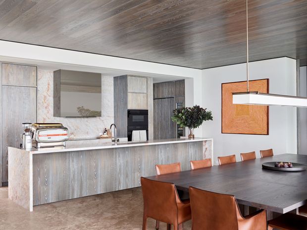 Contemporary open-plan kitchen and dining area featuring a long stone island with integrated sink, pale timber-look cabinetry, built-in appliances, a dark timber dining table, and leather dining chairs. The space has a dramatic dark timber ceiling, marble-look wall surfaces, warm neutral flooring, sculptural pendant lighting, and a restrained modern palette.