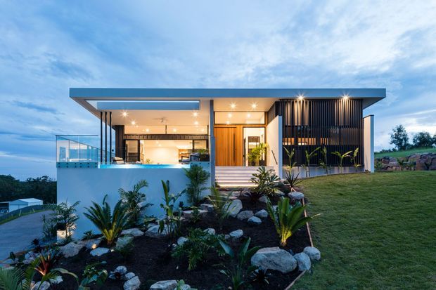 Contemporary single-storey house viewed from the landscaped front garden at dusk. The home has a broad flat roof with deep overhangs, extensive floor-to-ceiling glazing, warm timber-clad entry elements, and a striking vertical black screening facade. Tropical planting, rocks, lawn, and a sloping site surround the elevated residence.