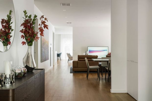 Bright open-plan contemporary interior viewed from an entry or hallway, with a dark timber console and decorative mirror in the foreground, leading into a living and dining area. The space features pale walls, timber flooring, a brown sectional sofa, dining table and chairs, extensive white built-in cabinetry, and soft filtered daylight through sheer curtains.